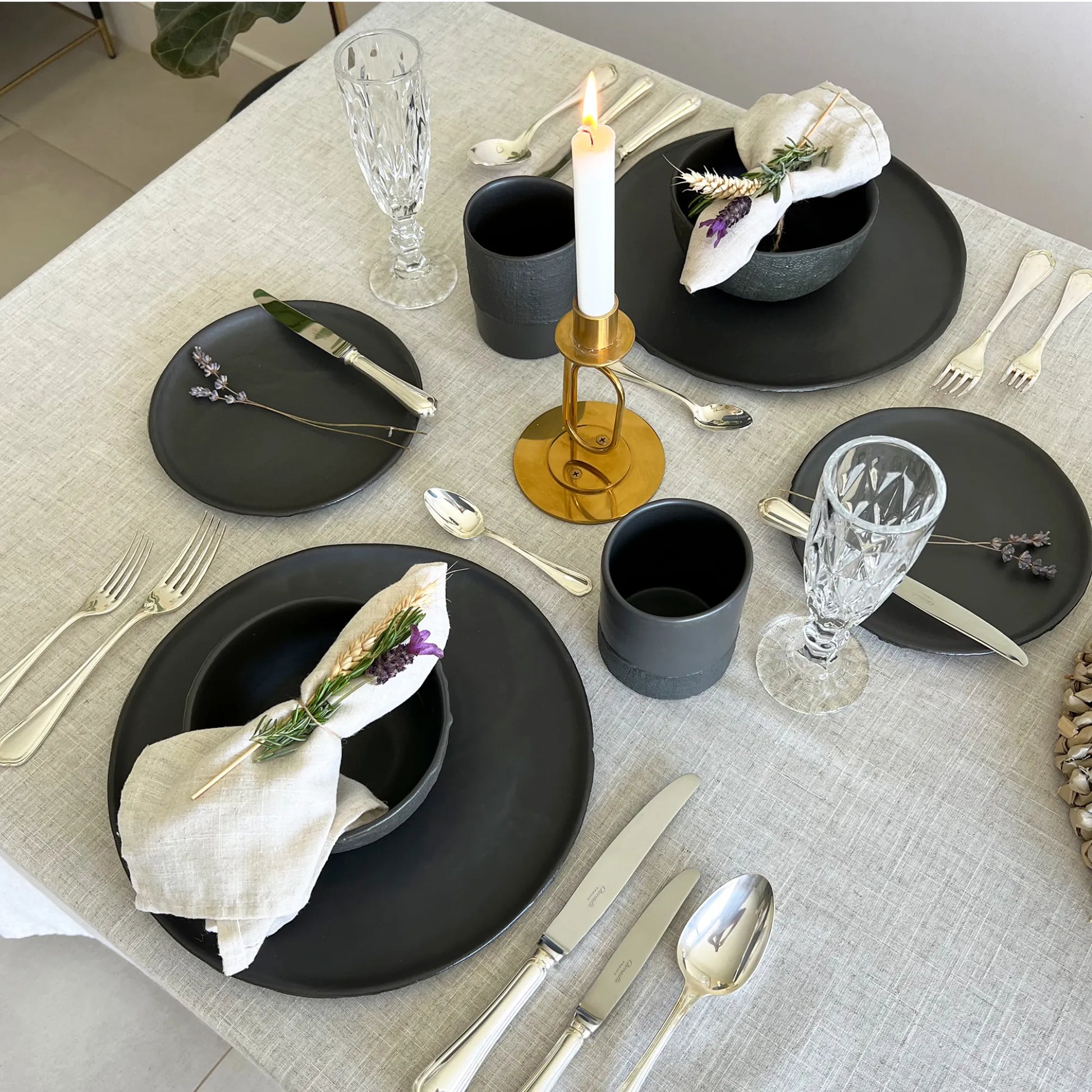 A styled dining table setting with black ceramic plates, matte black cups, black textured breakfast bowls, clear cut glass goblets, silver flatware, a gold candlestick with a white taper, and cream napkins with small purple floral accents, all arranged on a light beige linen tablecloth.