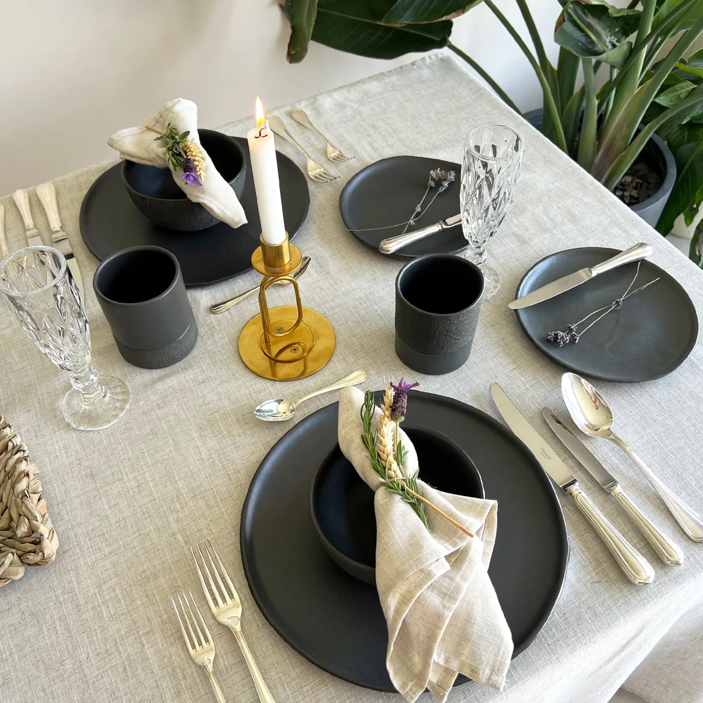 A styled dining table setting with matte black plates, dark gray cups, silver flatware, clear glassware, a gold candle holder with a lit white candle, and beige napkins with small purple floral accents, photographed in a bright indoor setting with a plant in the background.