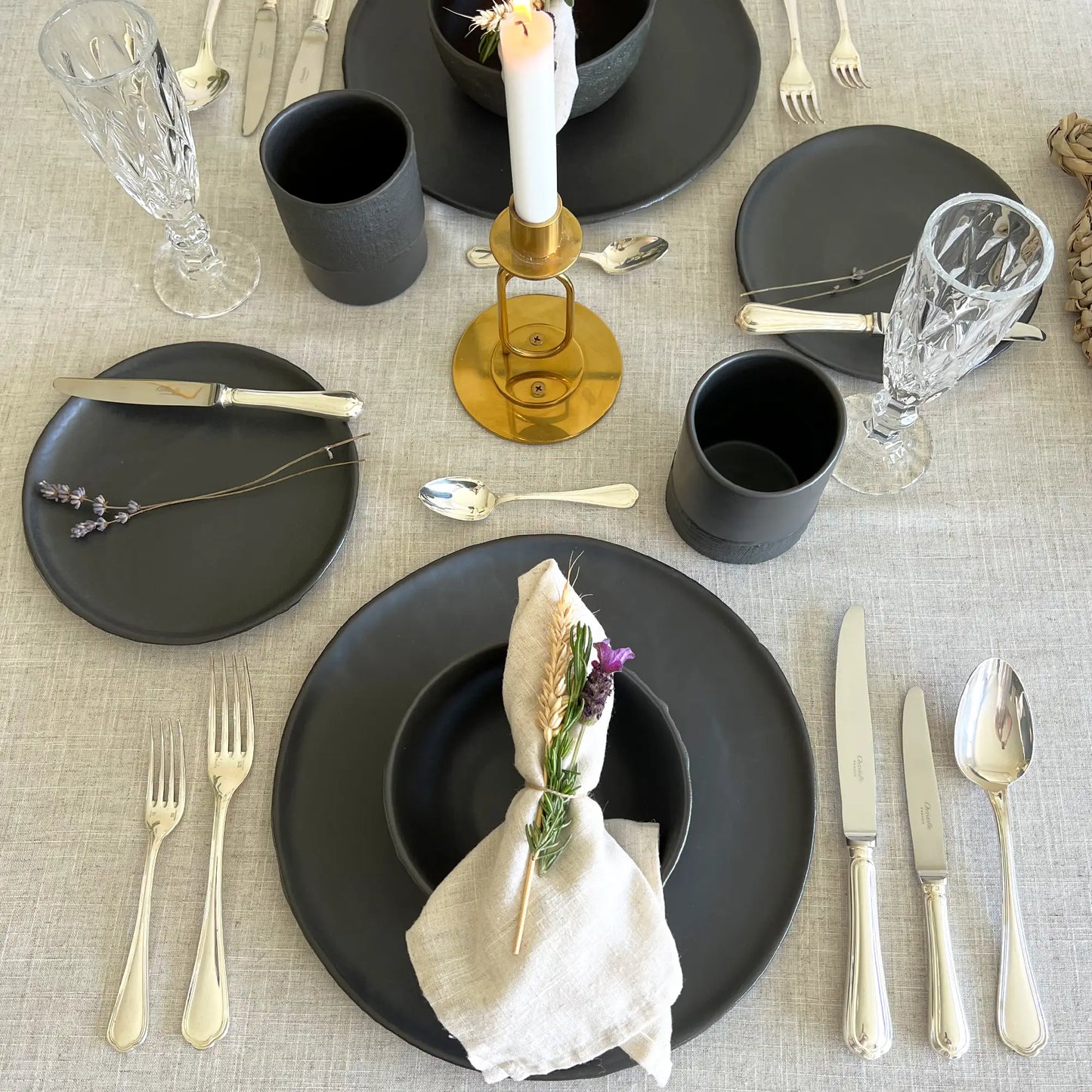 A styled tabletop place setting on a light beige linen tablecloth, featuring matte black ceramic plates and cups, silver flatware, clear crystal glasses, a gold candle holder with a lit white candle, and a cream napkin with a small purple flower accent.