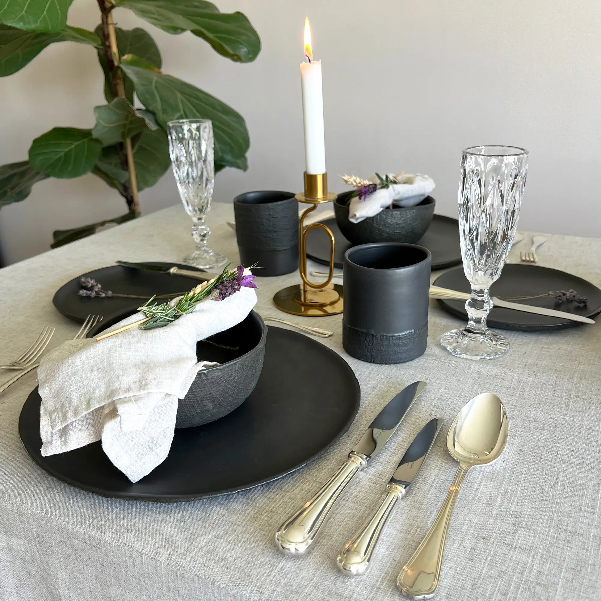 A styled dining table setting with matte black plates, black ceramic mugs, black soup bowls, silver flatware, clear cut-glass goblets, a white napkin, and a lit white candle on a light beige linen tablecloth.