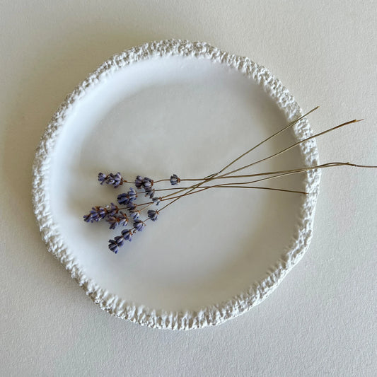A round dessert or side white ceramic plate with a textured, raw uneven rim, styled with a small sprig of dried lavender on a neutral beige background.