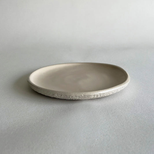 A dessert, shallow, round ceramic plate with a textured, hand-painted finish in a light warm grey or beige colour, set against a neutral light grey background.