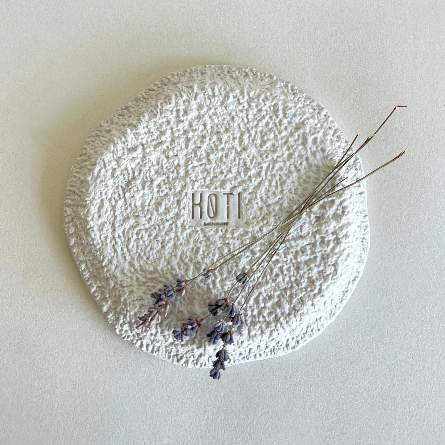 A textured round white side plate with a rough, uneven finish, featuring a central square brand logo 