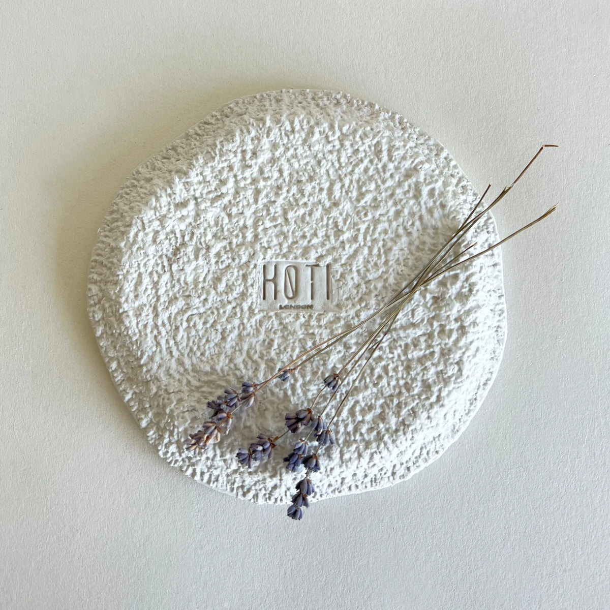 A textured round white side plate with a rough, uneven finish, featuring a central square brand logo 