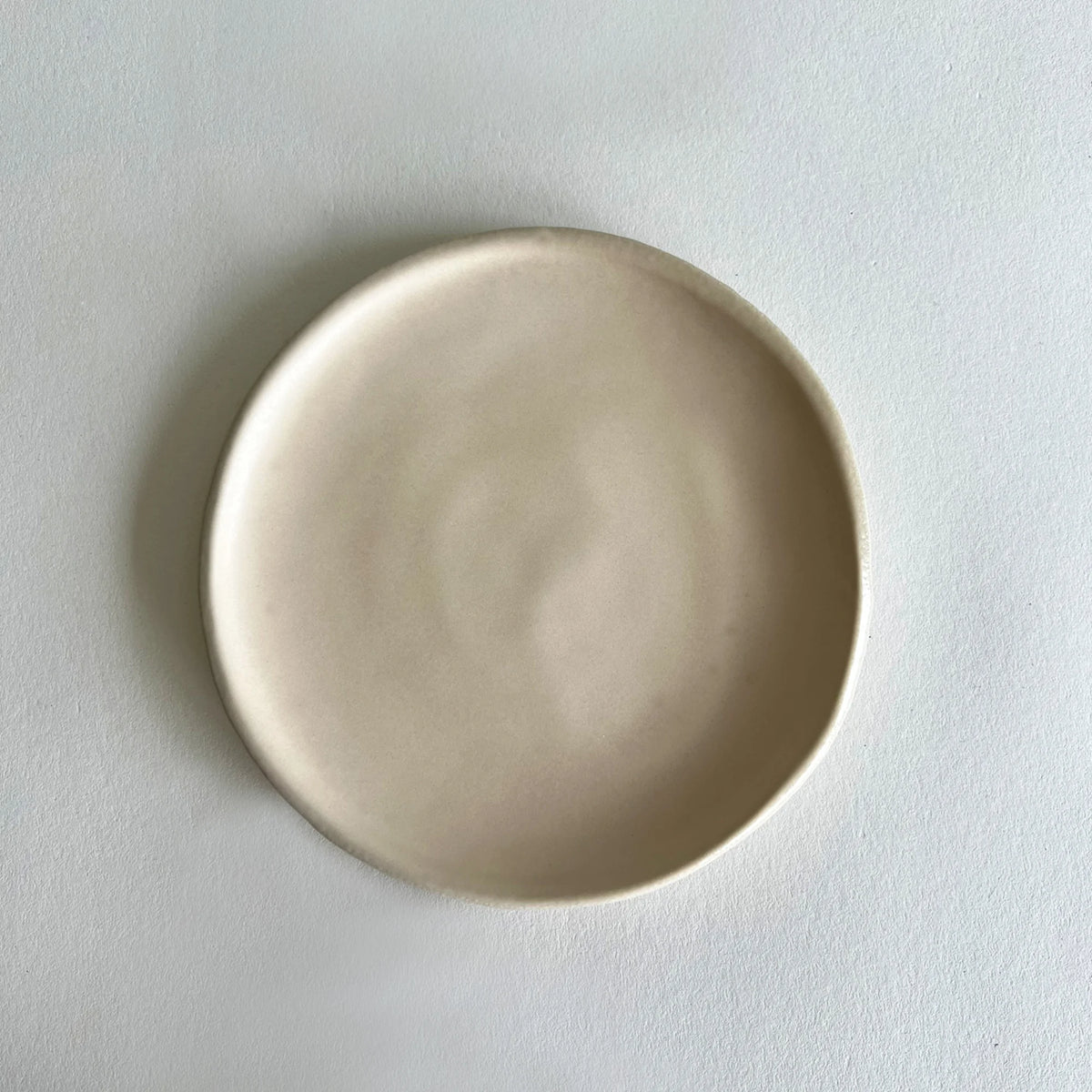 A minimalist, round ceramic plate in a matte, light beige finish, photographed from directly above on a textured white surface.