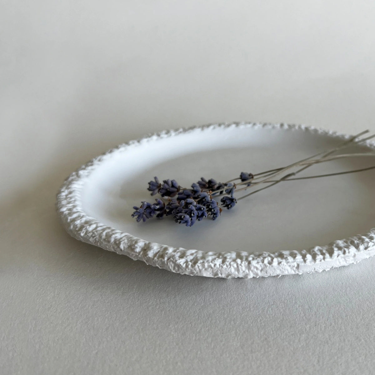 A white textured ceramic side plate with a raised, lacy-like rim, styled with dried lavender on a neutral light gray background.