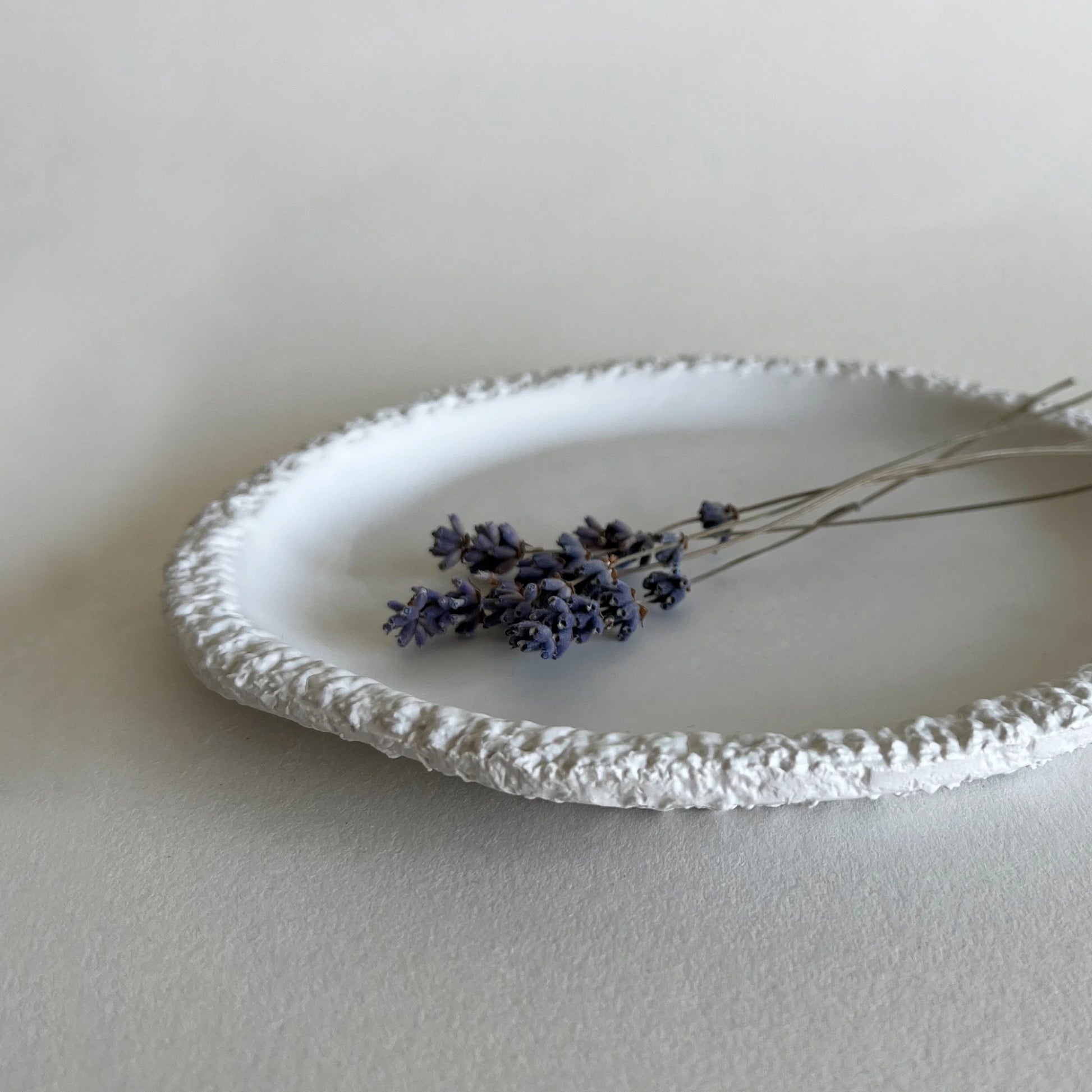 A white textured ceramic side plate with a raised, lacy-like rim, styled with dried lavender on a neutral light gray background.