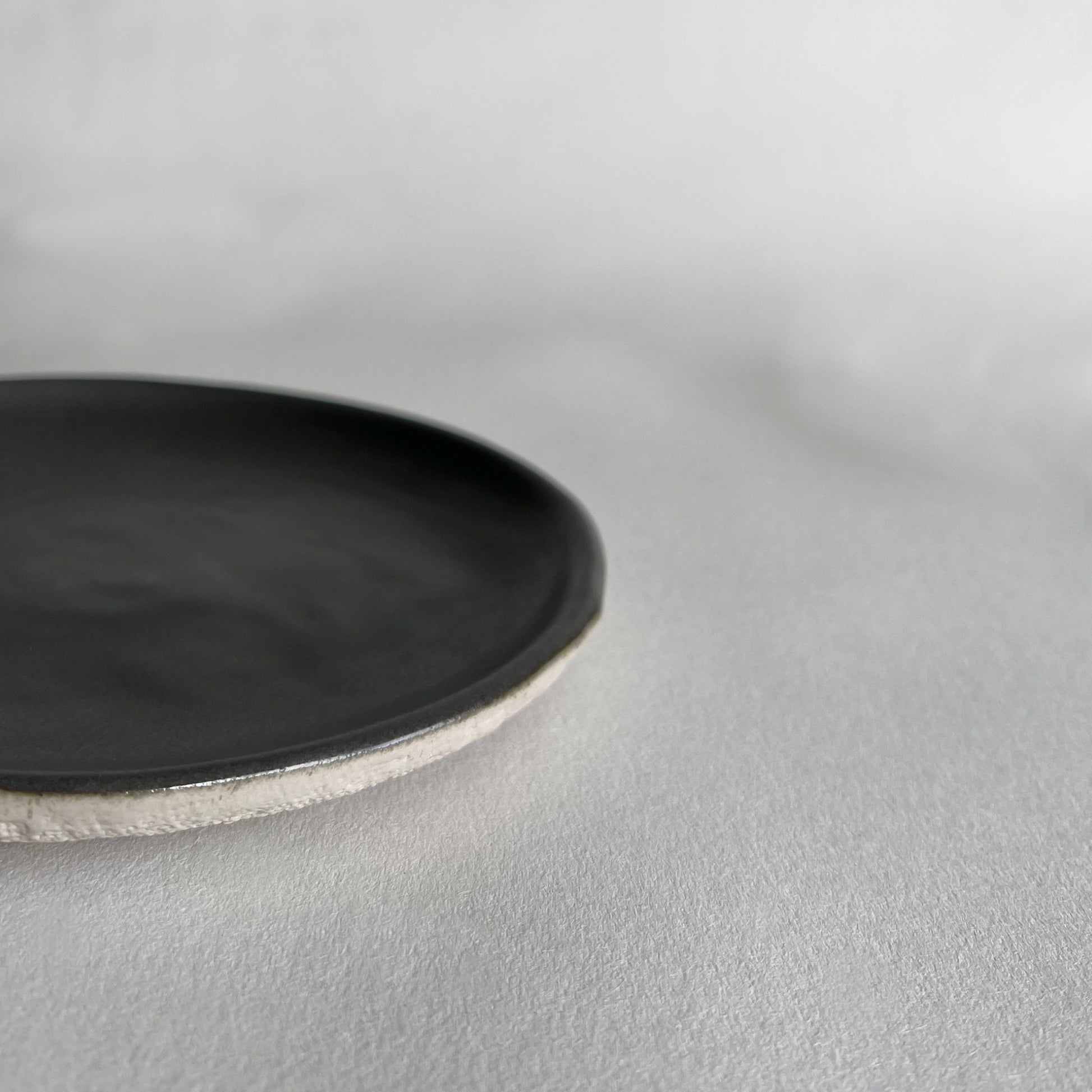Close up of handmade ceramic side plate in dark grey colour in matte finish, contrasted with beige organic texture.