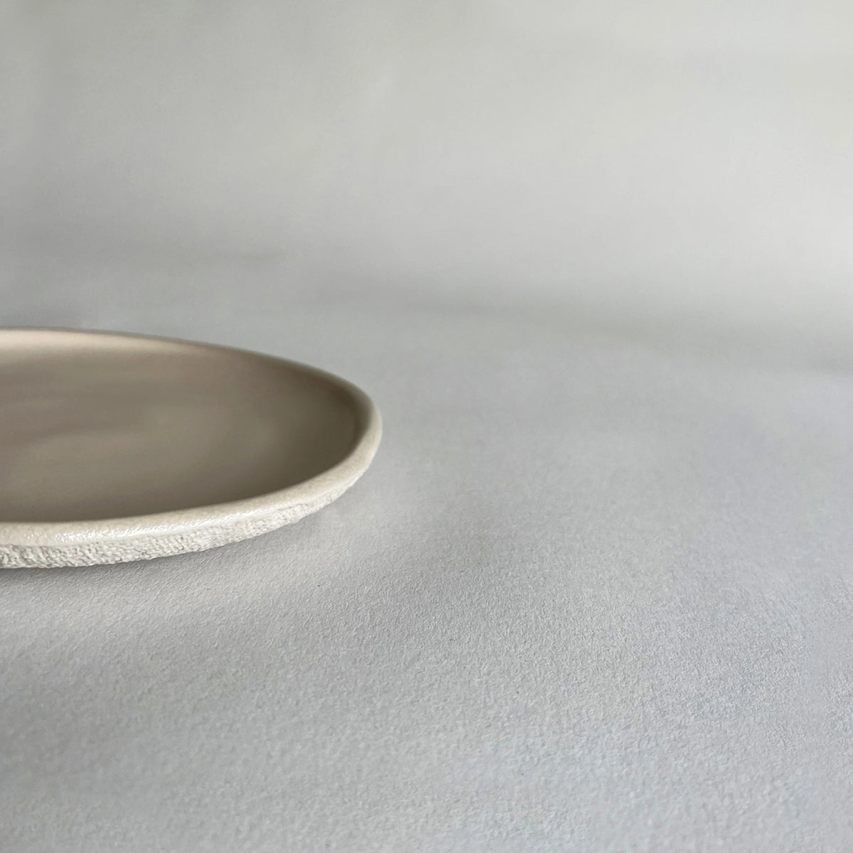 A close-up of a round ceramic dessert plate with a textured, matte finish in a light warm beige colour, set against a matching light grey background.