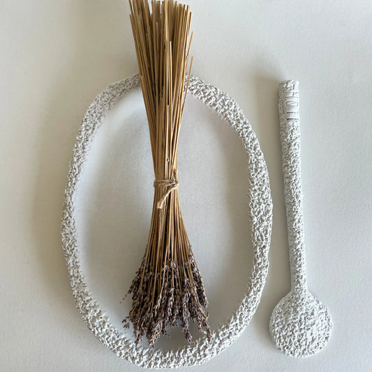 A textured white ceramic serving platter with a dried lavender sprig placed inside it, alongside a matching white textured serving spoon.