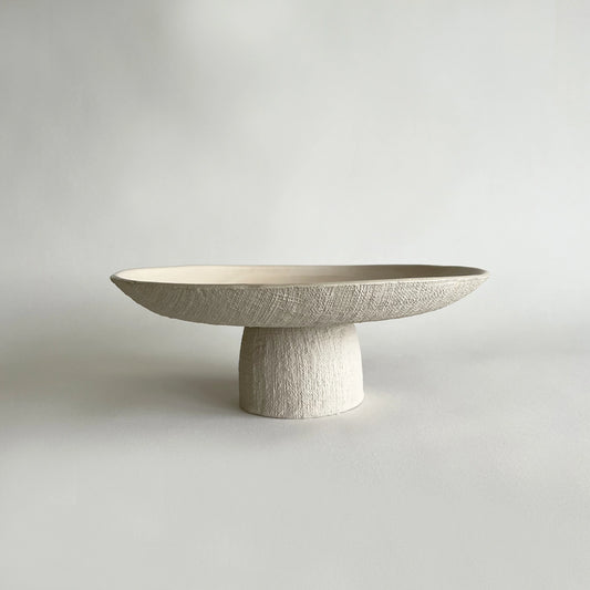 A minimalist, low-profile handmade ceramic pedestal tray with a wide, shallow oval top and a textured cylindrical base, finished in a warm off-white/cream tone with a subtle linen-like texture, set against a neutral light grey background.