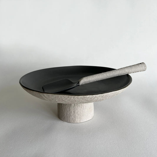 Beige and charcoal ceramic serving platter with a textured light beige pedestal base and a matching matte serving spoon.