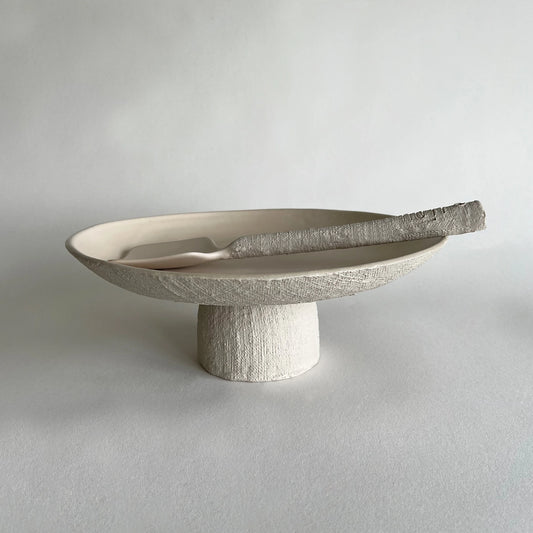 A textured ceramic pedestal tray and serving platter in a warm beige tone with a matching ceramic serving spoon, set against a neutral light grey background.
