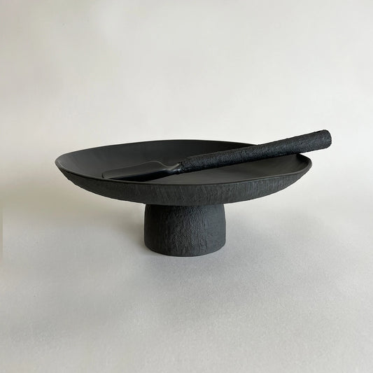 A matte black textured ceramic pedestal platter with a matching serving spatula, showcasing organic linen finish.