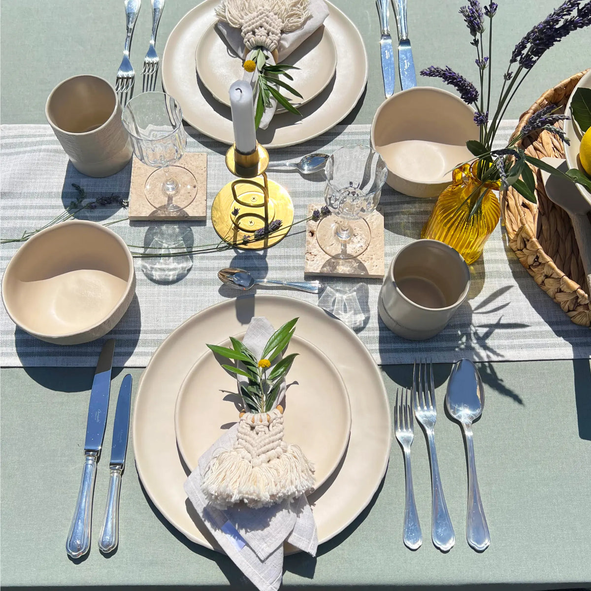 An outdoor table setting displayed with beige matte textured ceramic dinner plates, side plates, soup and breakfast bowls, coffee mug, using sage linen table cloth and linen napkins, finished with organic decoration such as olive tree branches.