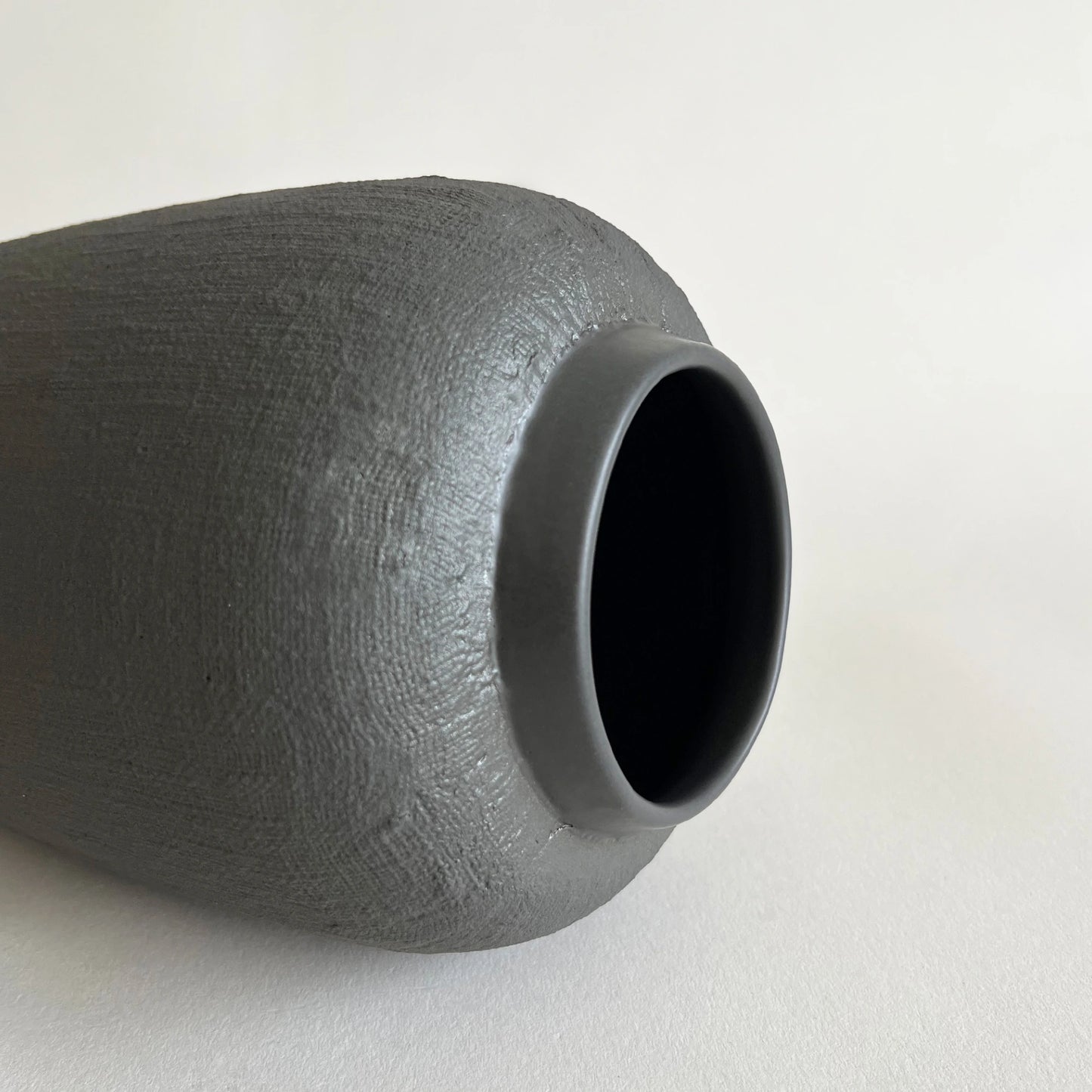 A close-up of a dark grey textured ceramic vase with a matte finish and a wide flared rim, close up of linen jute organic finish.