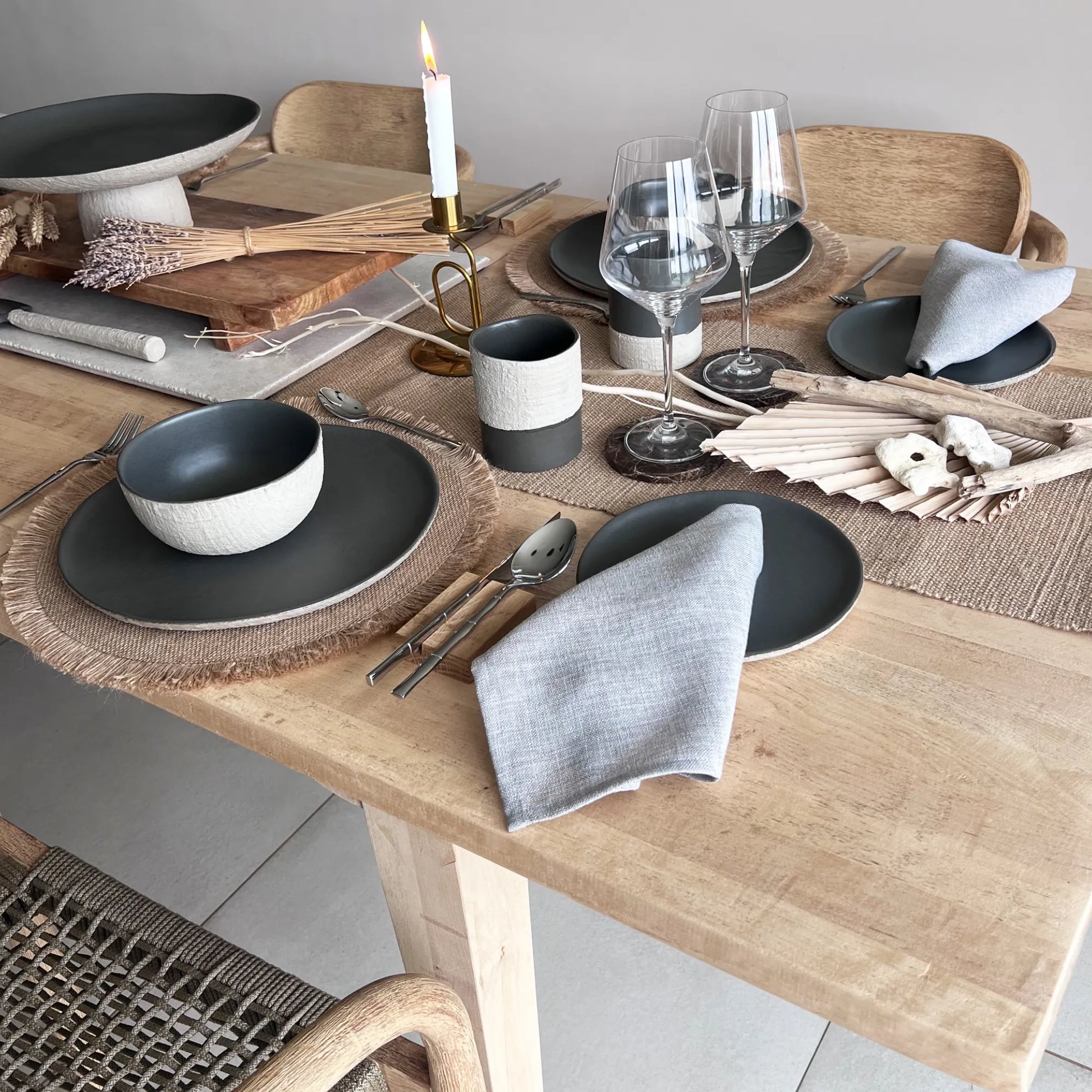 Dining table set with matte dark grey plates, charcoal bowls, and textured cups on a wooden table, showcasing rustic style.