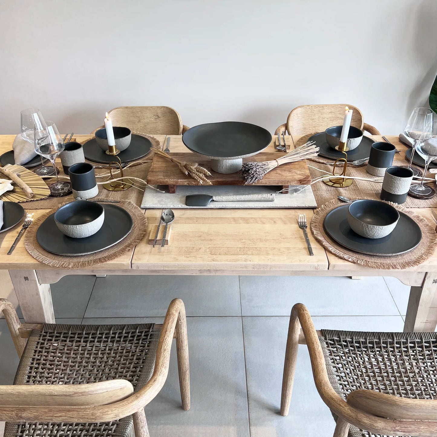 Dining table set with beige and charcoal plates, bowls, cups and matching pedestal serving platter, and cutlery on a wooden table with wicker chairs.