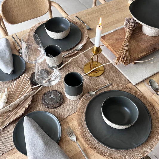 Dining table setting with beige and charcoal colour dinner plates, salad plates, matching bowls and coffee cups, grey napkins, and a candle on a wooden table.
