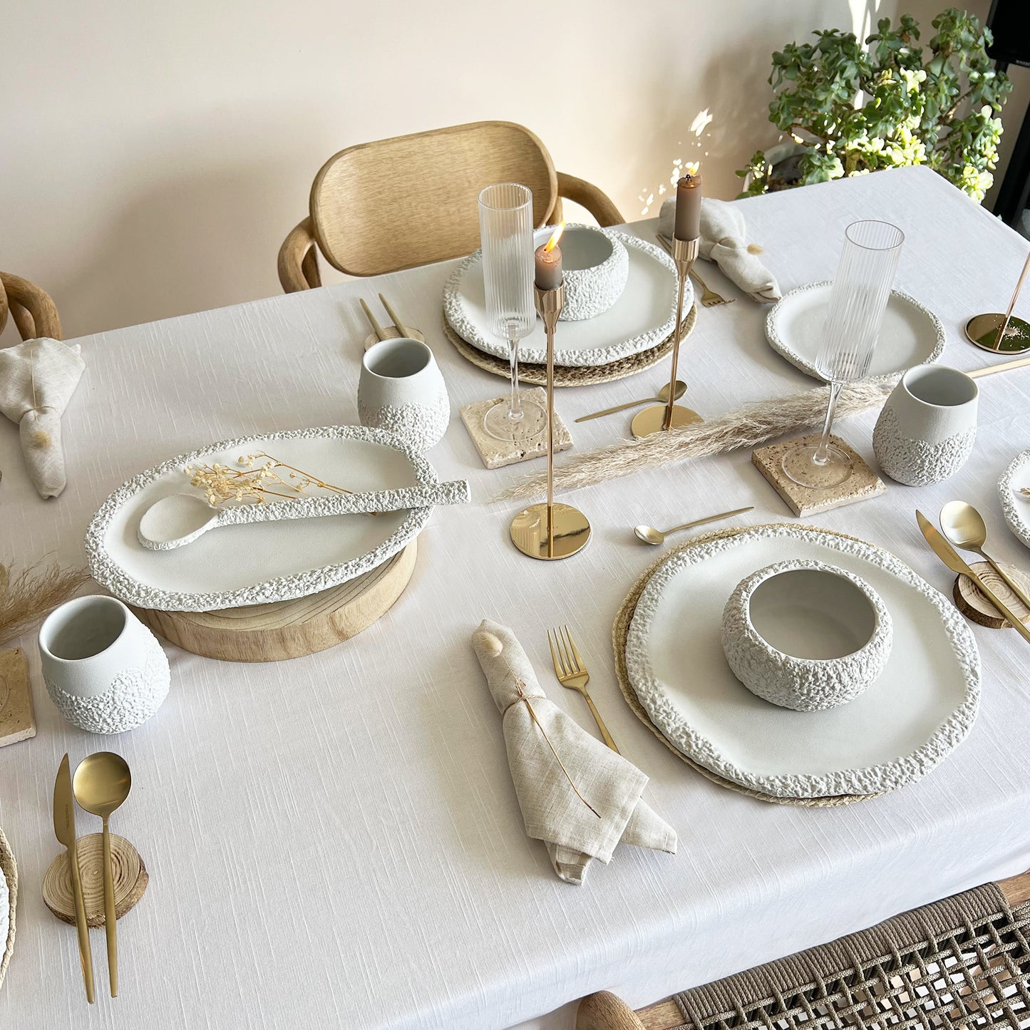 Elegant table setting with white handmade ceramic dinnerware set, gold cutlery, and decorative elements on a light-coloured tablecloth.