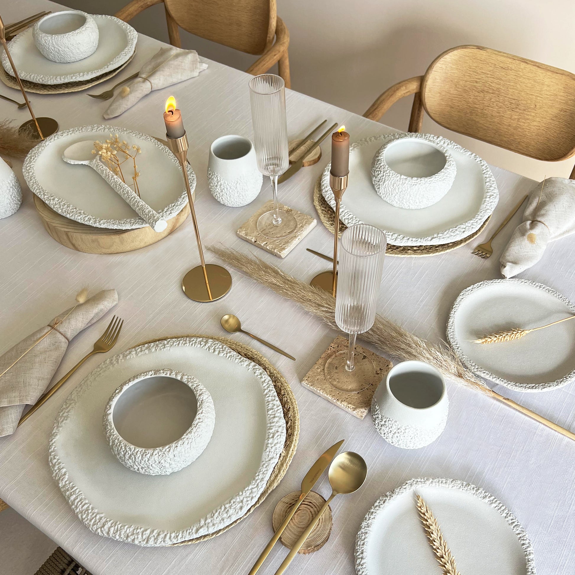 Elegant table setting with white ceramic plates, bowls and cups, gold cutlery, and candles on a white tablecloth.