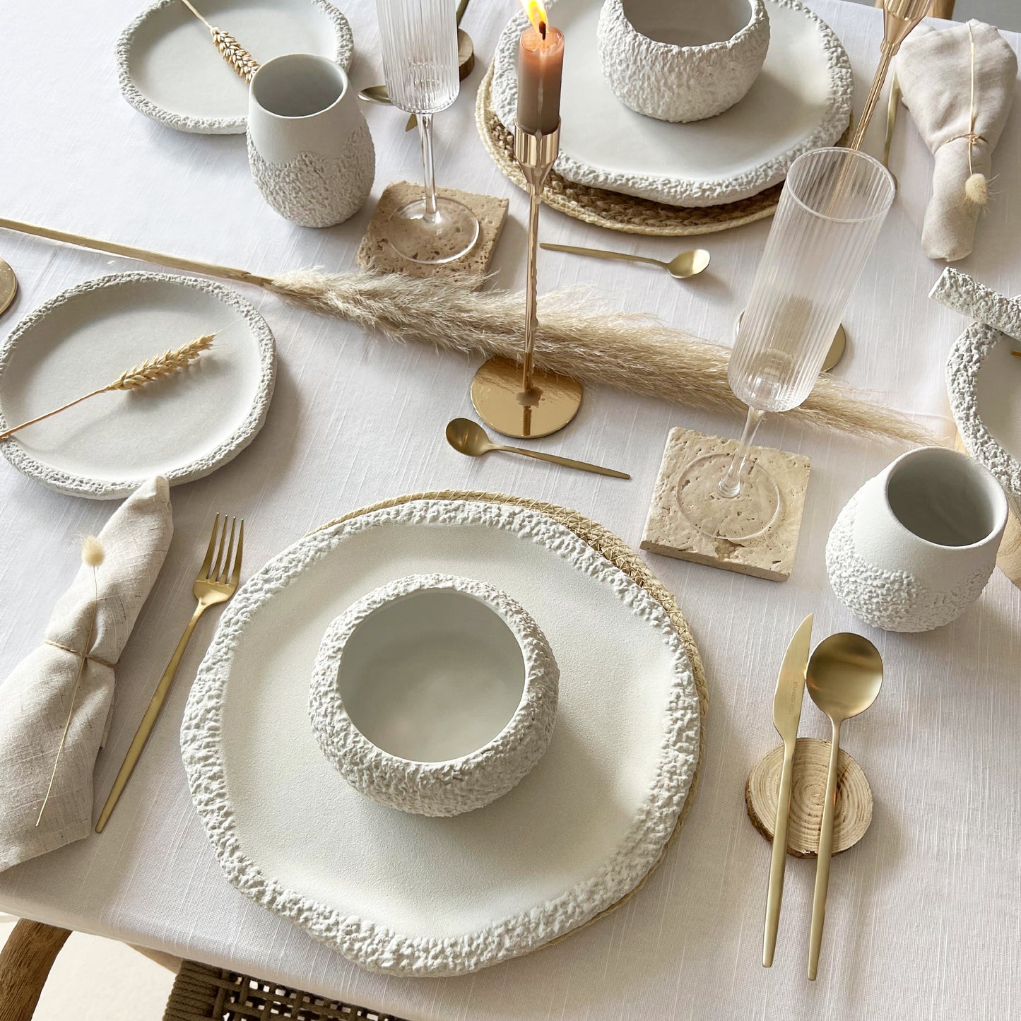 Elegant table setting with white ceramic dinnerware set, gold cutlery, and beige decorative elements on a light background.