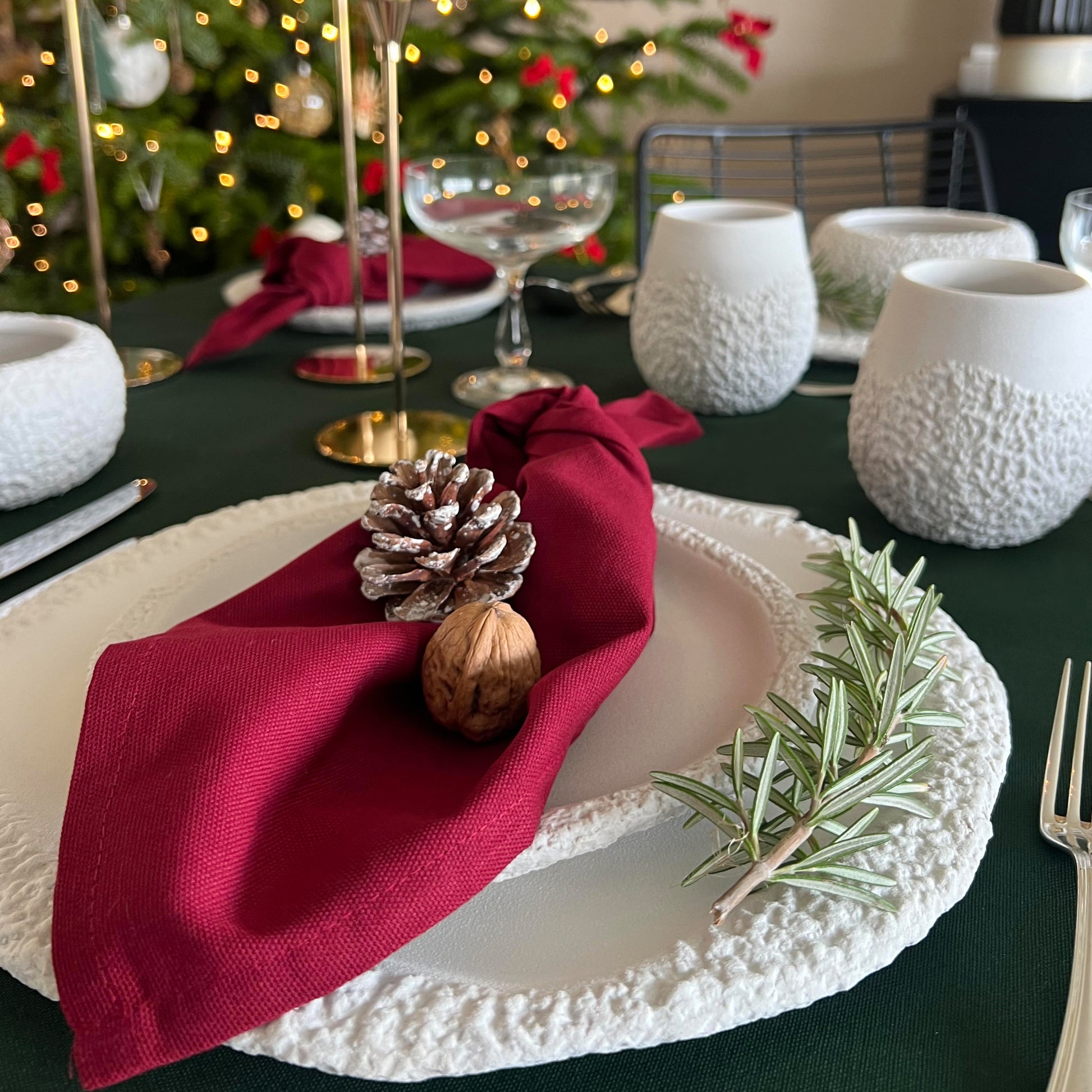 Decorative table setting with red napkin, pine cone, and rosemary on a white ceramic dinnerware set