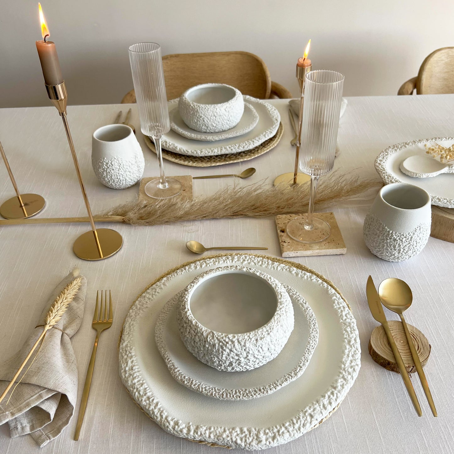wedding table setting with white ceramic plates, bowls and cups, gold cutlery, and candles on a light surface.