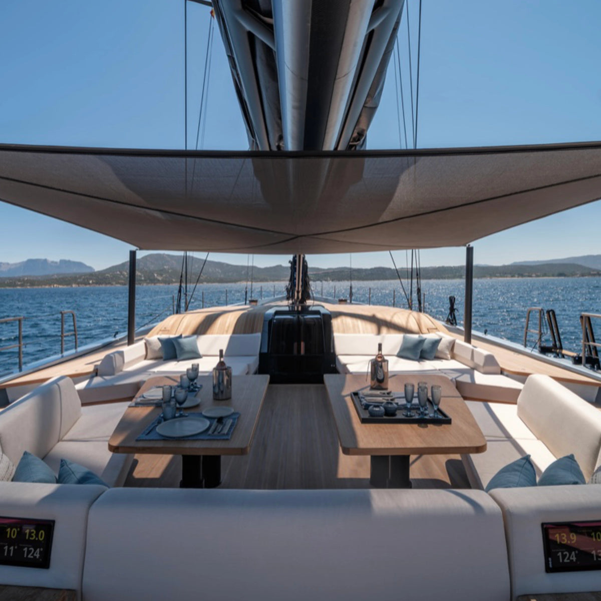 A luxury yacht deck featuring a spacious outdoor seating area with cream cushions, two wooden dining tables set with glassware, ceramic plates and cups, and a large beige bimseal canopy overhead, with blue ocean water and distant hills under a clear sky.