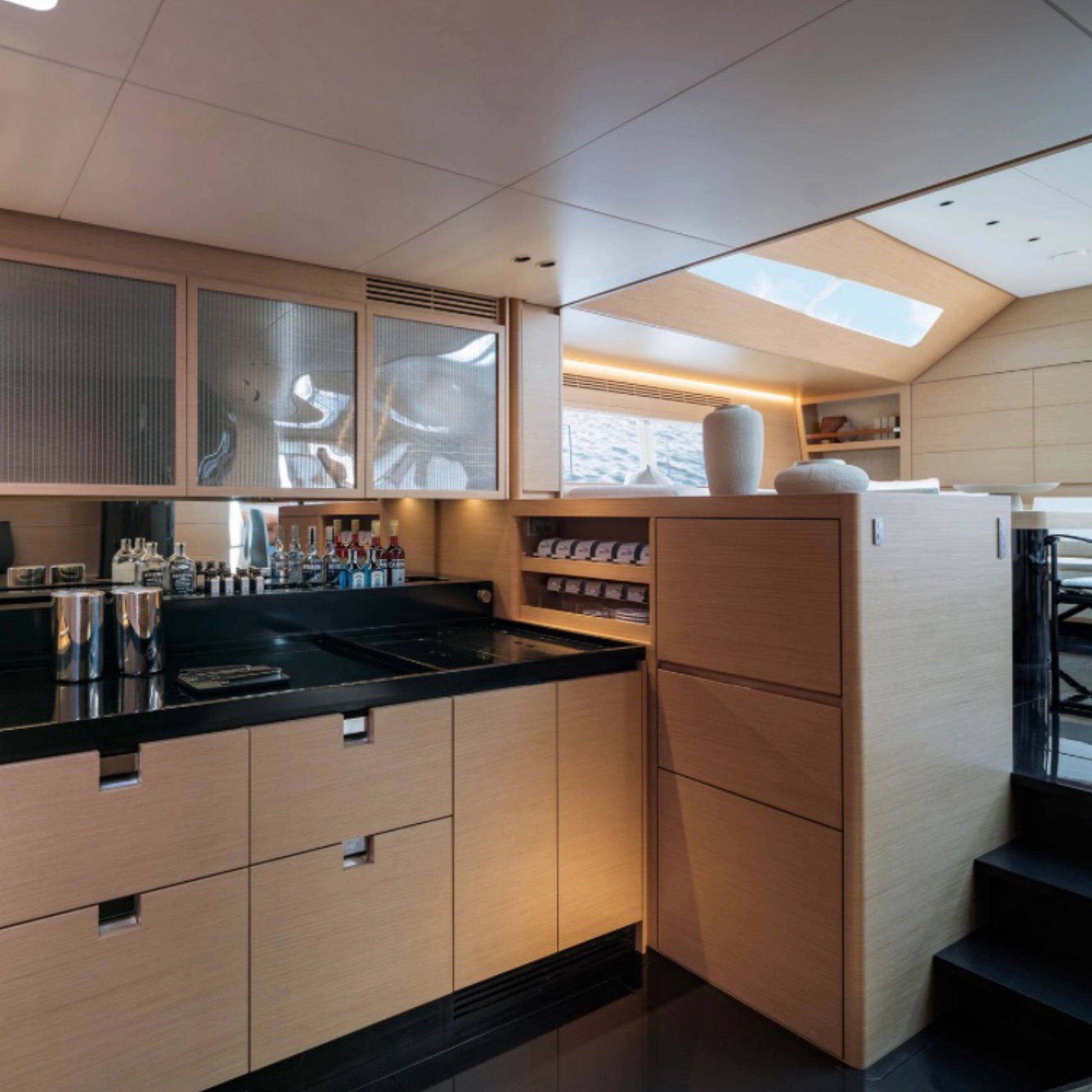 Modern yacht galley kitchen interior with light wood cabinetry, decorated with tall and smaller beige textured vases, black countertops, glass-front upper cabinets, and a staircase on the right.