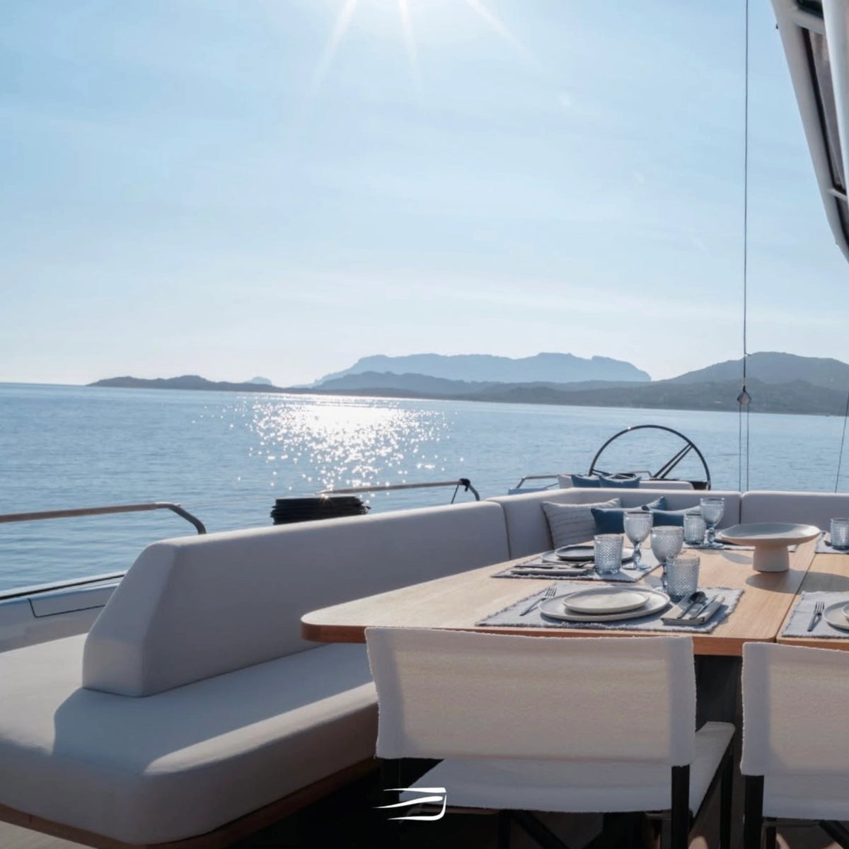 Outdoor dining area on a yacht  with a wooden table set with ceramic plates and glasses, white upholstered seating, and a scenic ocean view with distant mountains under a bright sun.