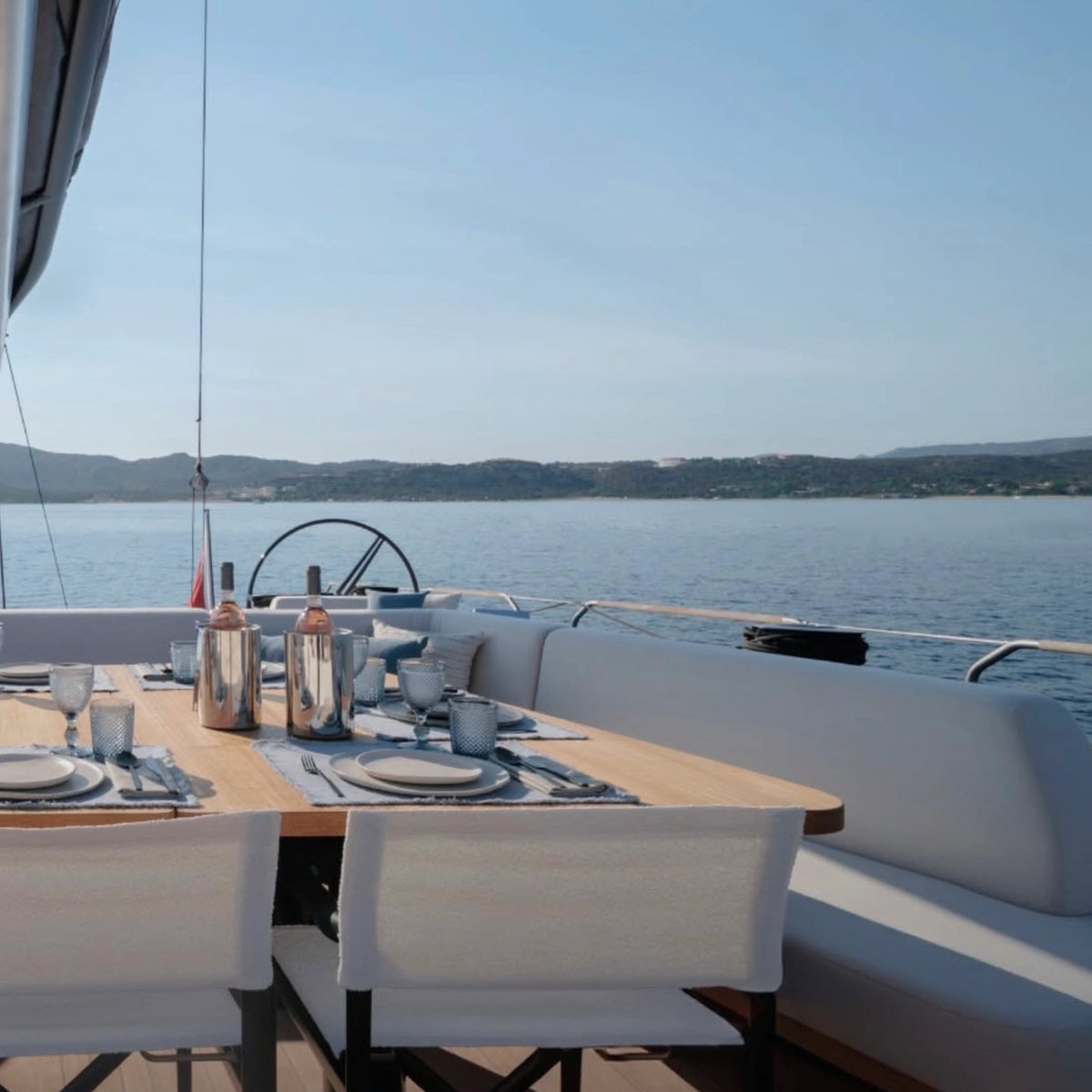 A luxury outdoor dining area on a yacht , featuring a wooden table set with plates and cutlery, white upholstered chairs, and a white cushioned bench overlooking a calm blue sea under a clear sky.