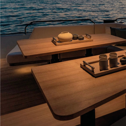 A modern wooden table set on a yacht  with ocean water in the background. The table has a warm light-wood finish and is styled with minimalist ceramic vessels and ceramic coffee cups.