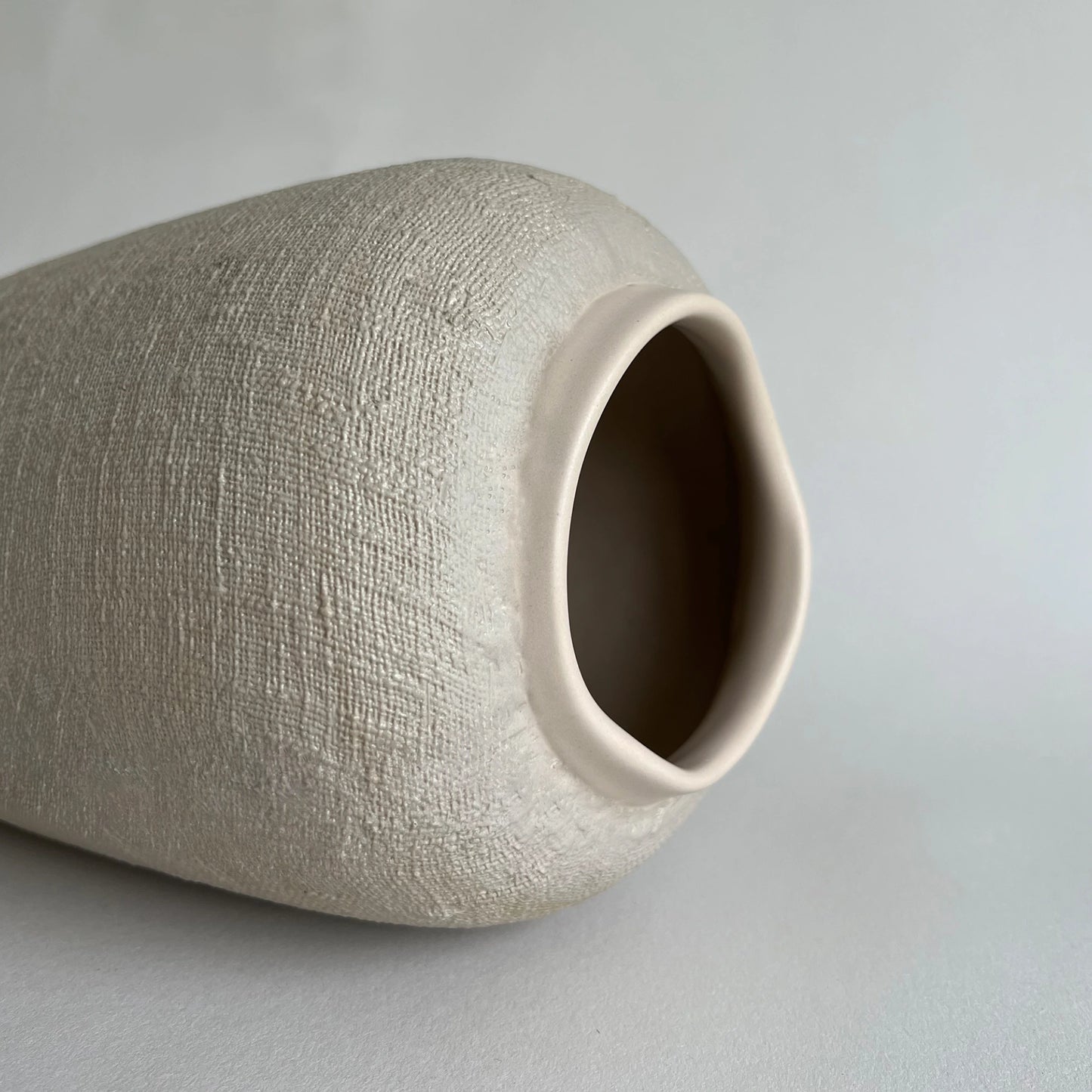 A close-up of a textured ceramic vase with a matte, linen-like beige finish and a small flared rim, photographed on a neutral light gray background.
