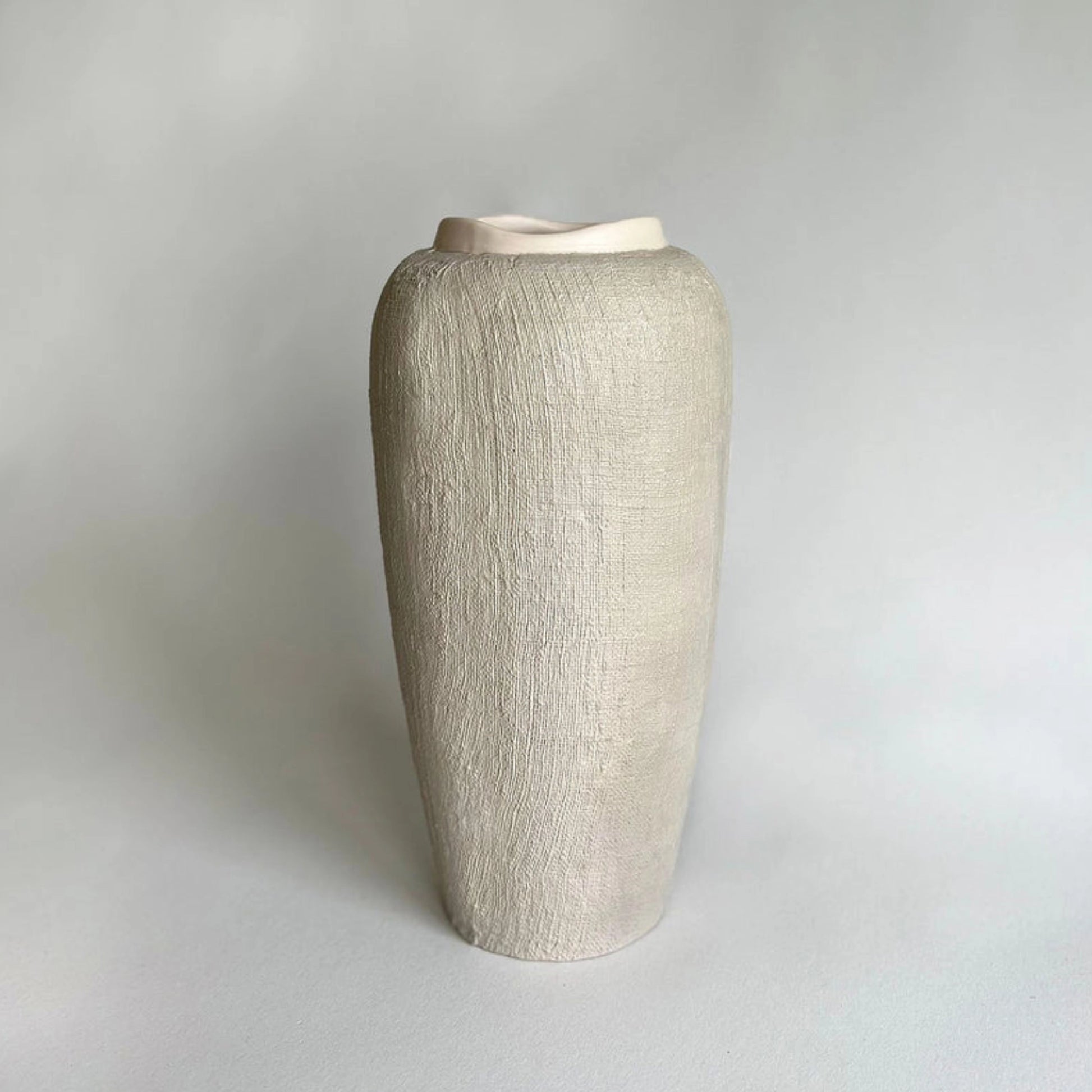A tall, beige handmade textured ceramic vase with a flared rim, with organic linen texture.