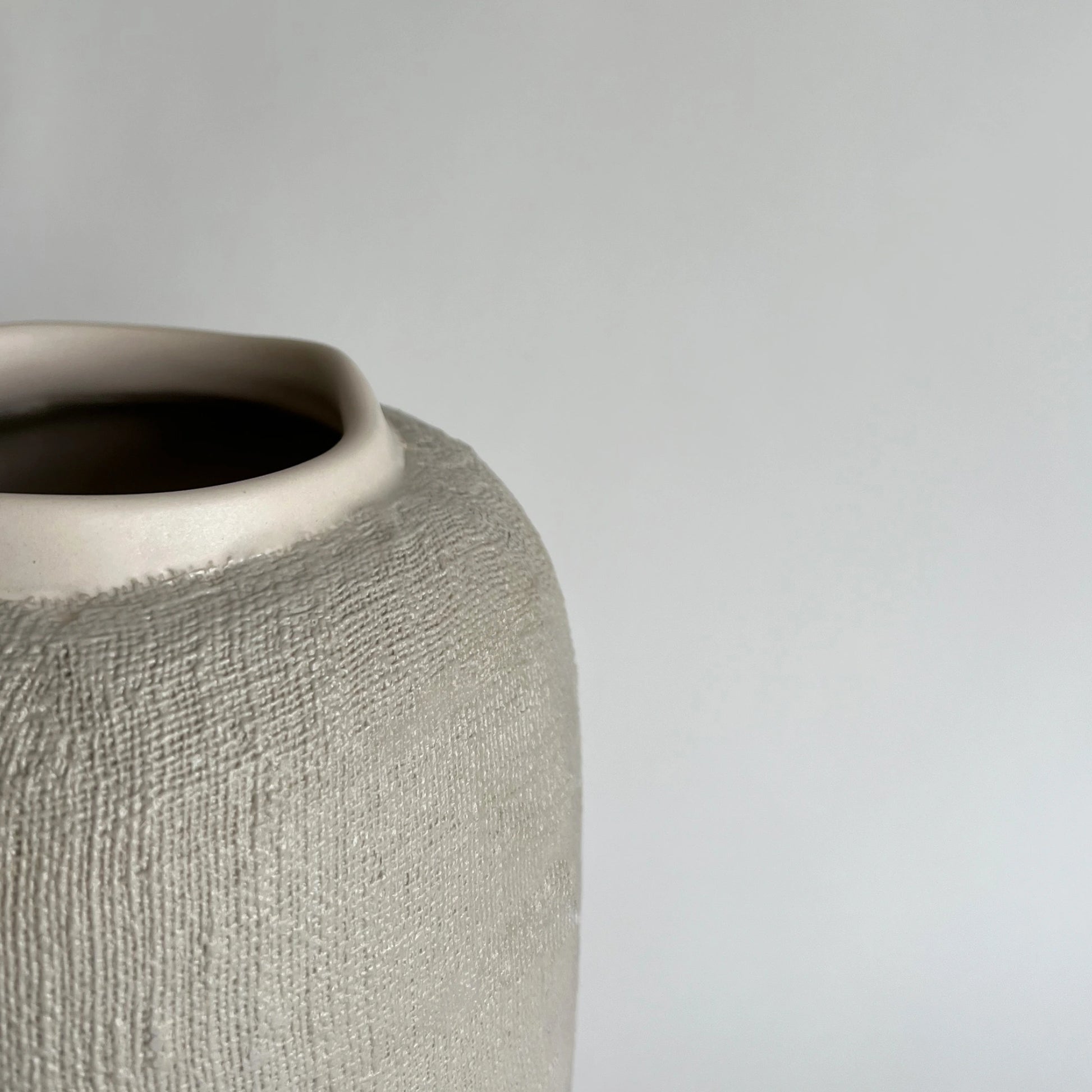 Close-up of a large textured ceramic vase in a warm light beige or greige tone, showing the side profile and open top against a plain light grey background.