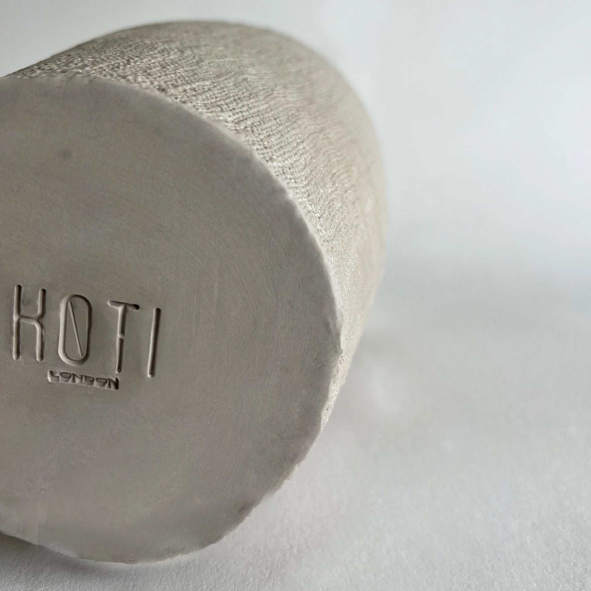 A close-up of a cylindrical ceramic vessel base with a textured matte finish, showing the embossed brand name.