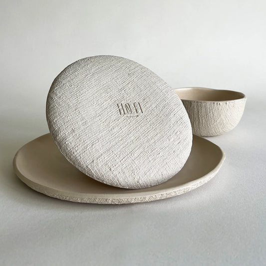 A minimalist ceramic table setting featuring a round textured side plate, dinner plate, with a breakfast textured bowl in the background against a neutral light grey backdrop.